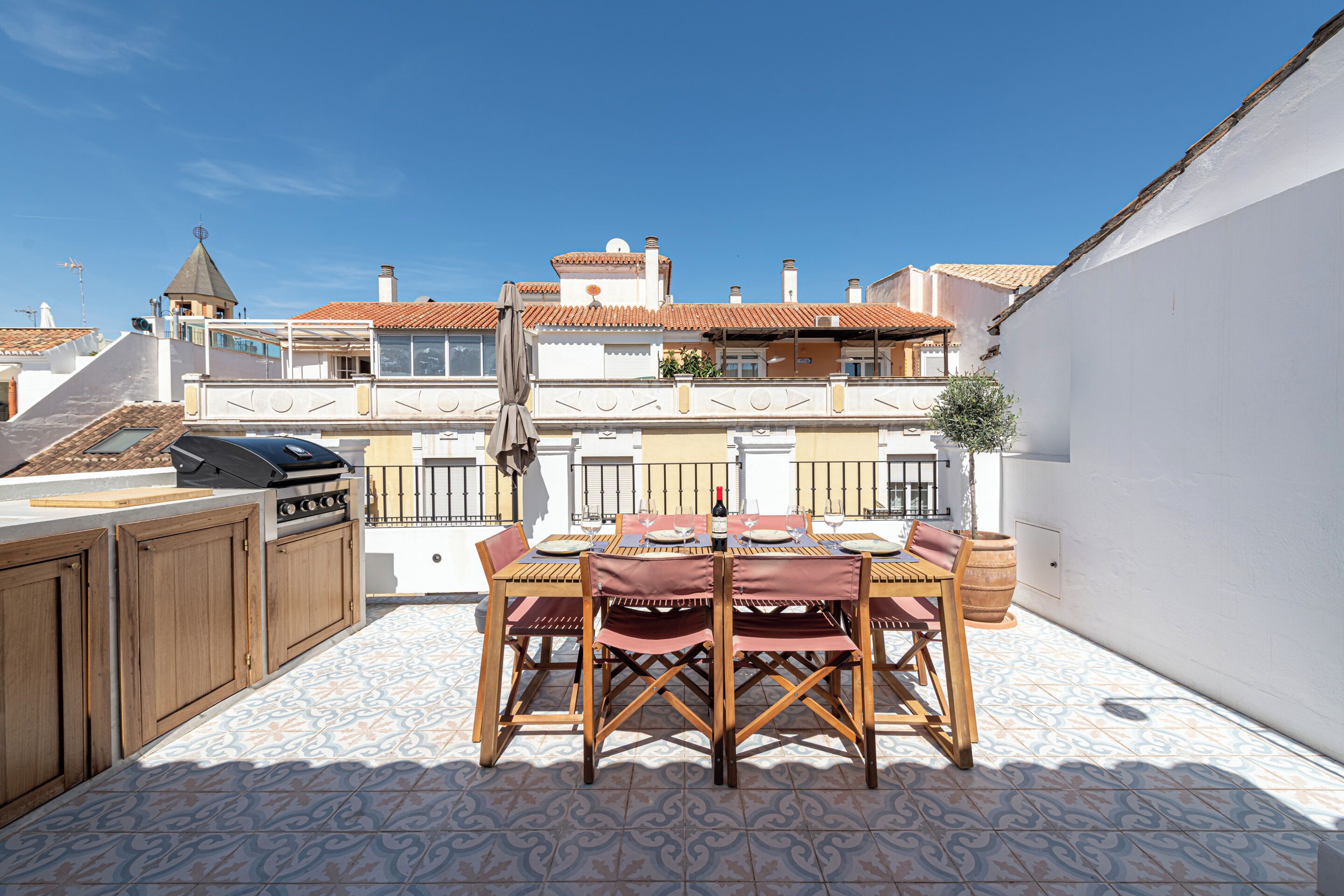 An outdoor patio with a dining table set for four, surrounded by wooden chairs and a barbecue grill.