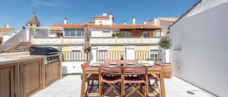 An outdoor patio with a dining table set for four, surrounded by wooden chairs and a barbecue grill.