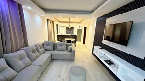 Ein modernes Wohnzimmer mit einer Eckcouch, einem runden Hocker, einem flachbildfernseher an der Wand und einem Essbereich im Hintergrund.