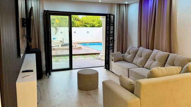 Ein modernes Wohnzimmer mit einem beigen Sofa, einem kleinen Hocker und einem Flachbildfernseher auf einem Holzboden. Eine Glastür öffnet sich zu einem Außenbereich mit einem Pool.