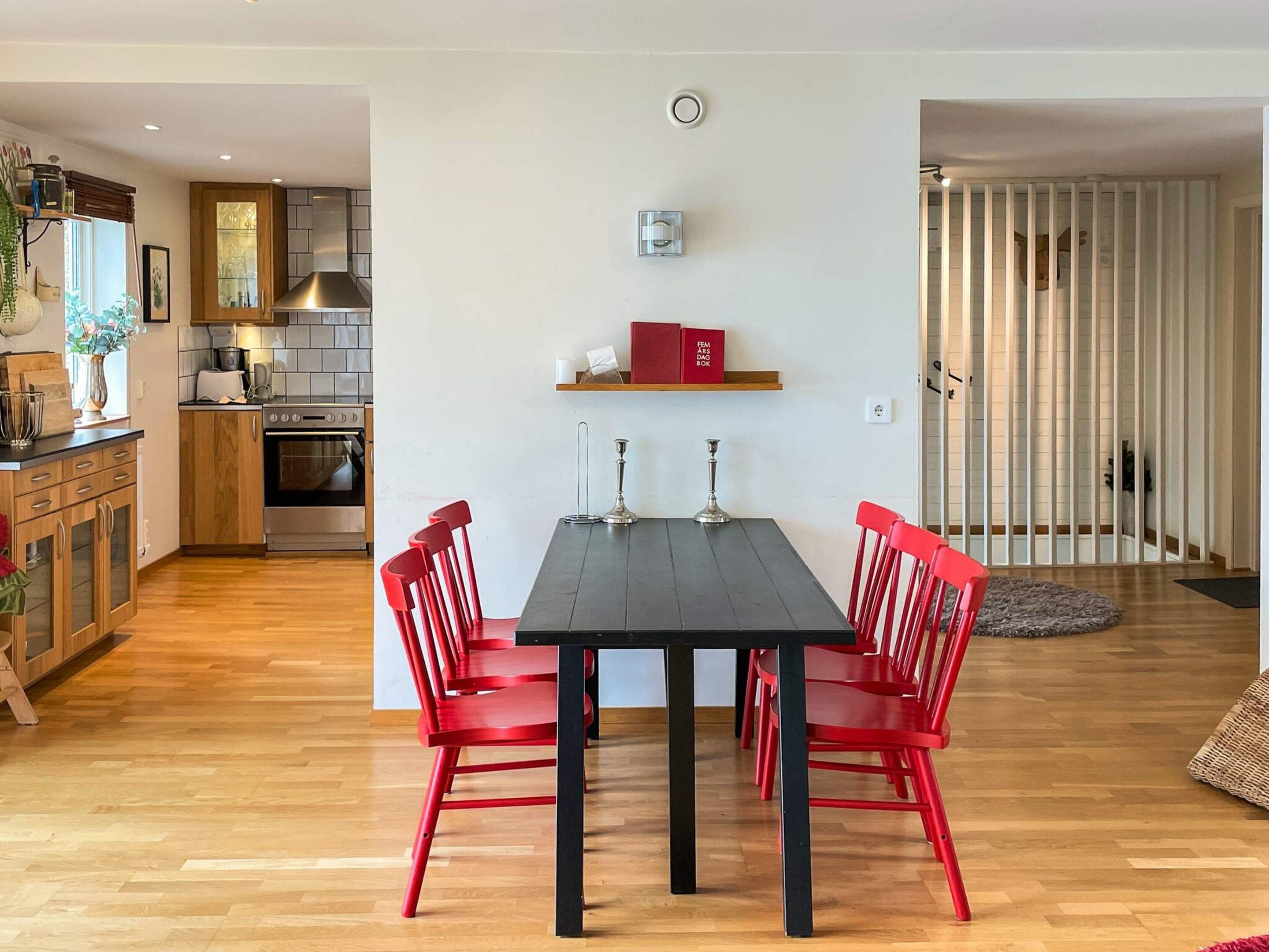 A modern kitchen with wooden cabinets, a built-in oven, and a dining area with a black table and red chairs.