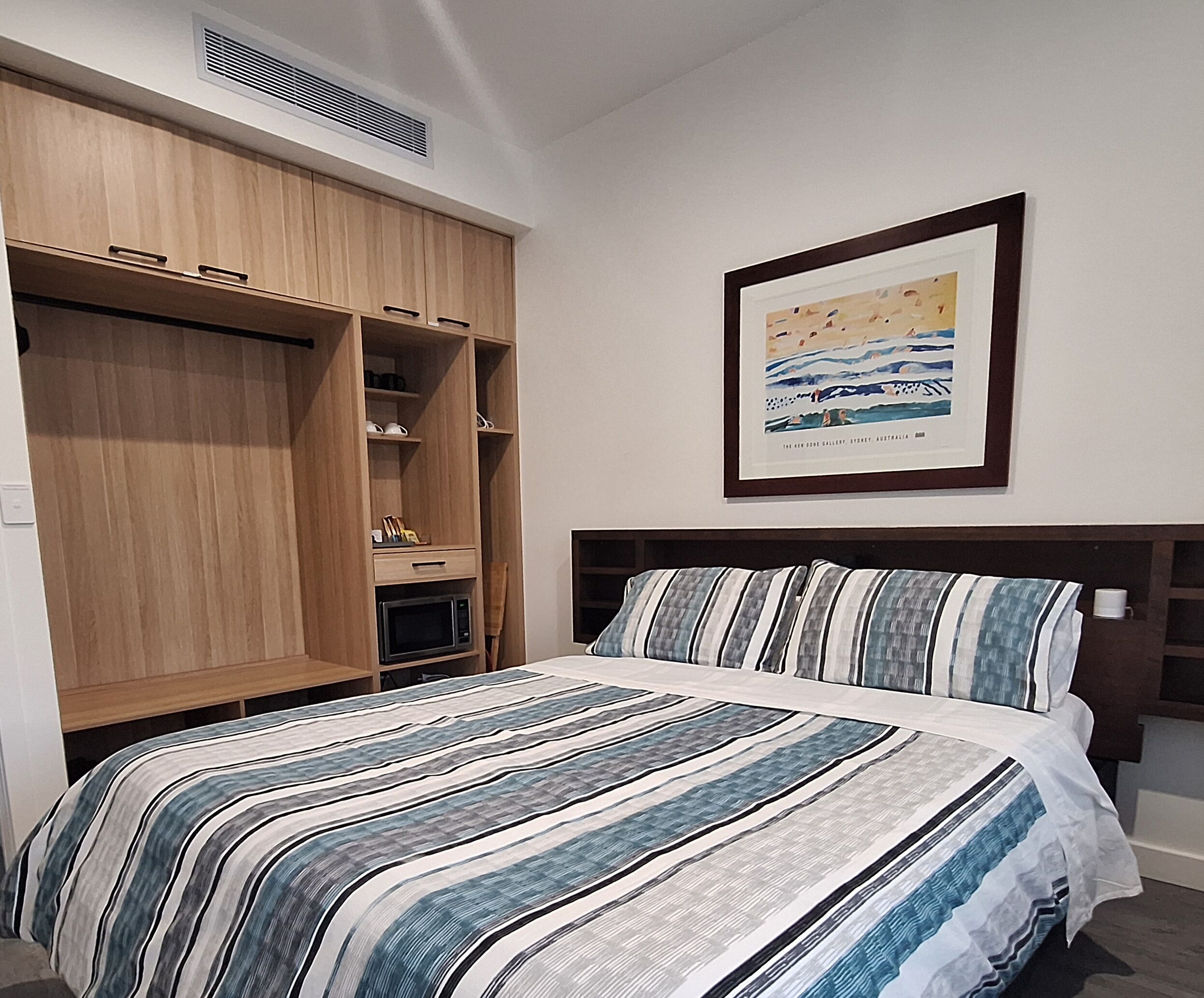 A bedroom with a bed, a wooden wardrobe, a framed picture, and a microwave.
