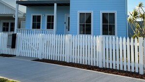 A two-story house with a white picket fence, a balcony on the upper floor, and a clear sky.