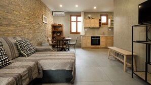 A living room with a brick wall, a sofa, a dining table with chairs, a kitchen area with wooden cabinets, and a TV on a stand. - Rianou Apartment (Chania)