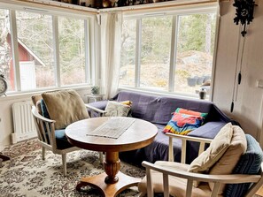 A cozy room with a round wooden table, a cushioned sofa, and a chair, with a view of trees and a building outside.