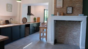 A modern kitchen with dark cabinets, a white countertop, and a brick wall. There is a fireplace and a hanging light fixture. - Le petit Santafé (Port-Sainte-Foy-et-Ponchapt)