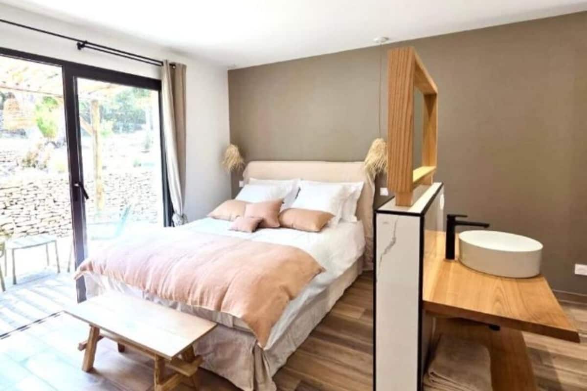 Un dormitorio moderno con una cama grande, un banco de madera, un estante empotrado con un espejo redondo y una mesita de noche pequeña con un lavamanos.