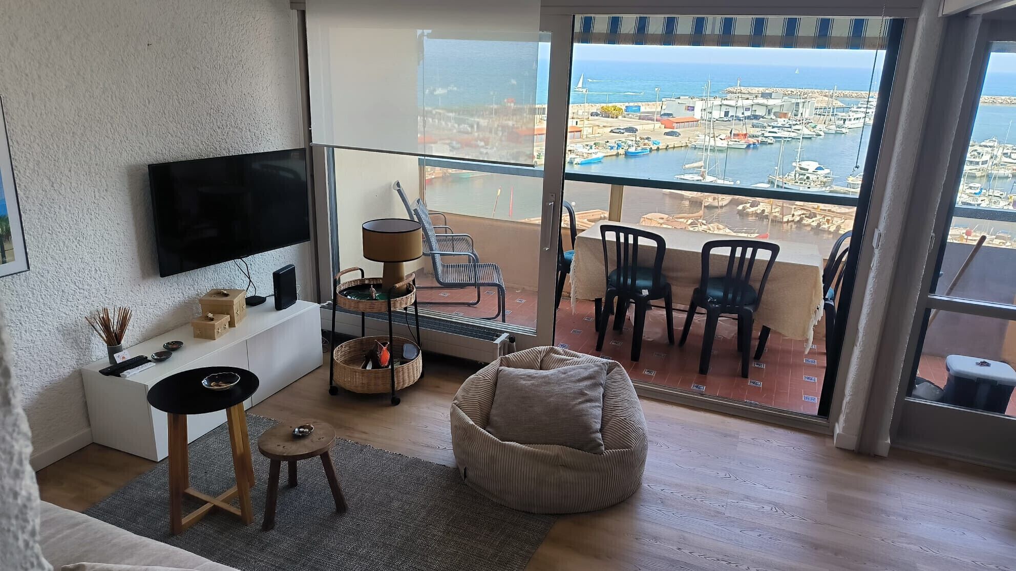 A modern living room with a large TV, comfortable seating, a small table, and a view of a marina through a glass door.