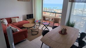 Een woonkamer met een rode bank, een televisie, een houten tafel en een balkon met rieten stoelen en uitzicht op zee.