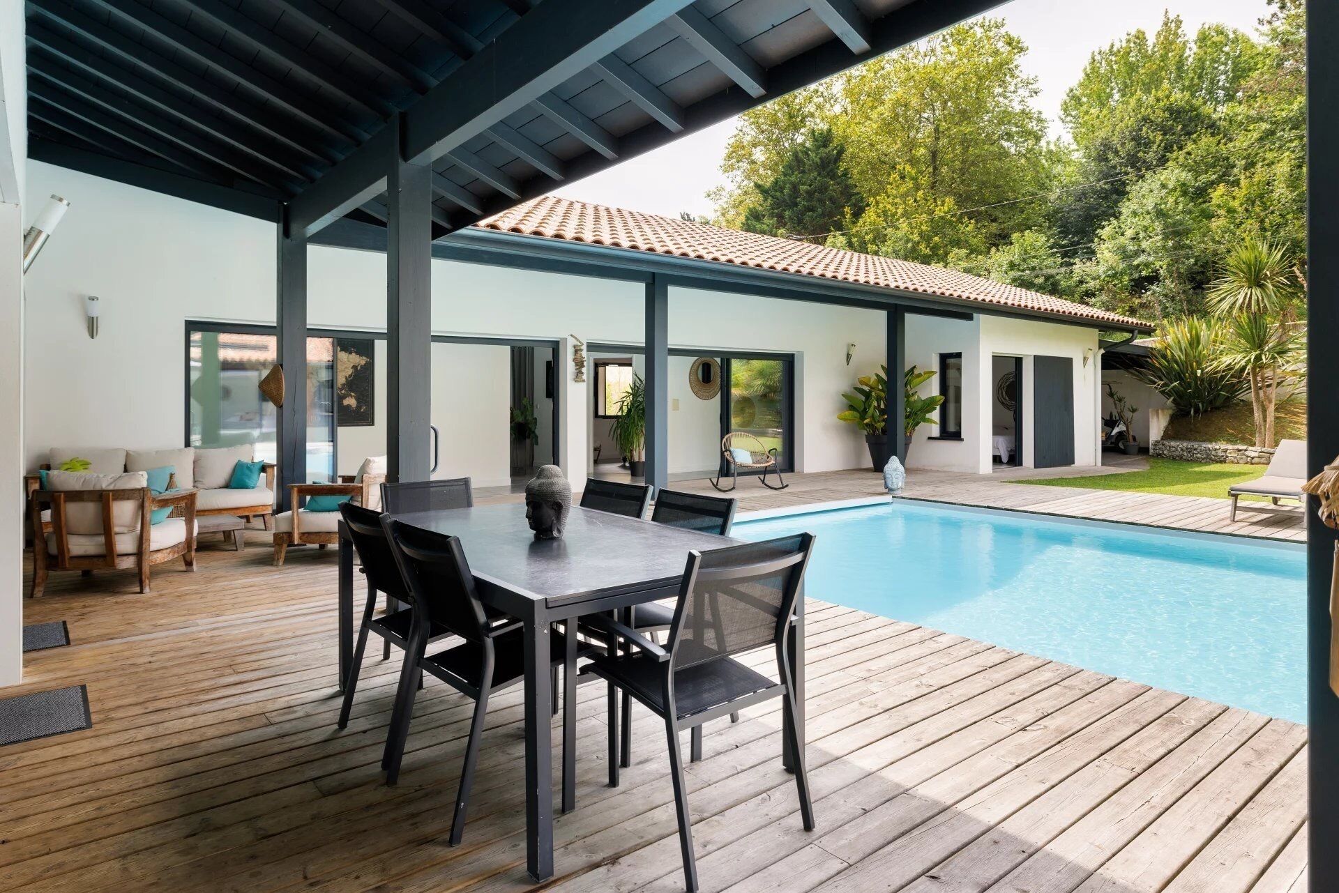 A modern outdoor pool area with a wooden deck, a dining table with chairs, and a covered seating area with a view of the pool.