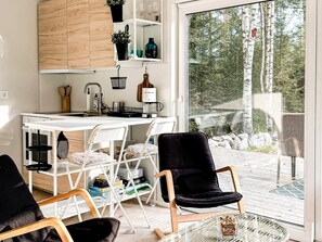 Una cocina moderna con armarios blancos, una zona de comedor con dos sillas negras y vista a un bosque a través de un ventanal.