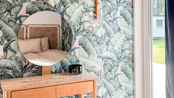 A bedroom with a round mirror, a bed with a woven headboard, a wooden nightstand, and a tropical wallpaper.