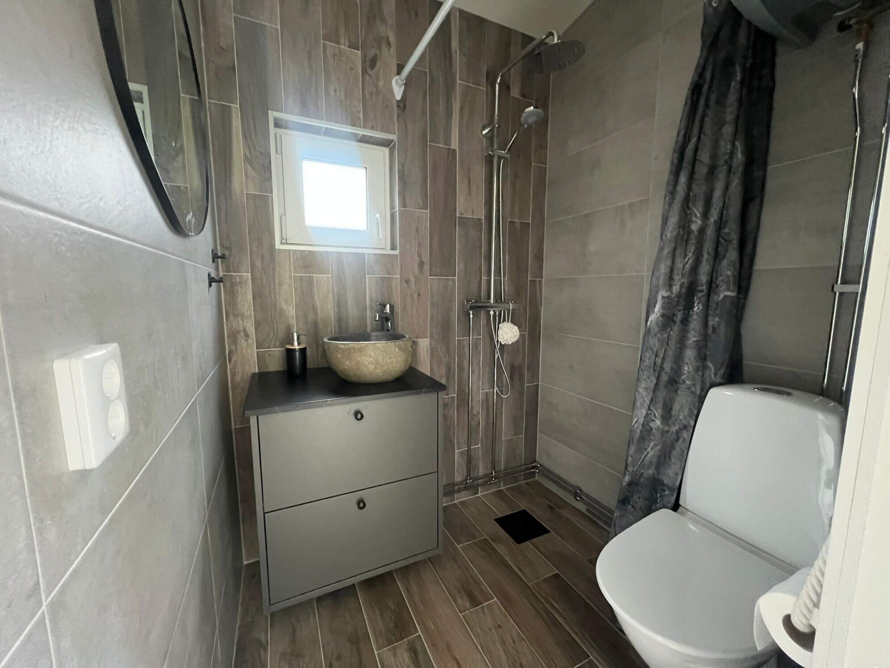 A modern bathroom with a wooden floor, a white toilet, a sink with a vessel basin, a mirror above the sink, and a shower with a curtain.