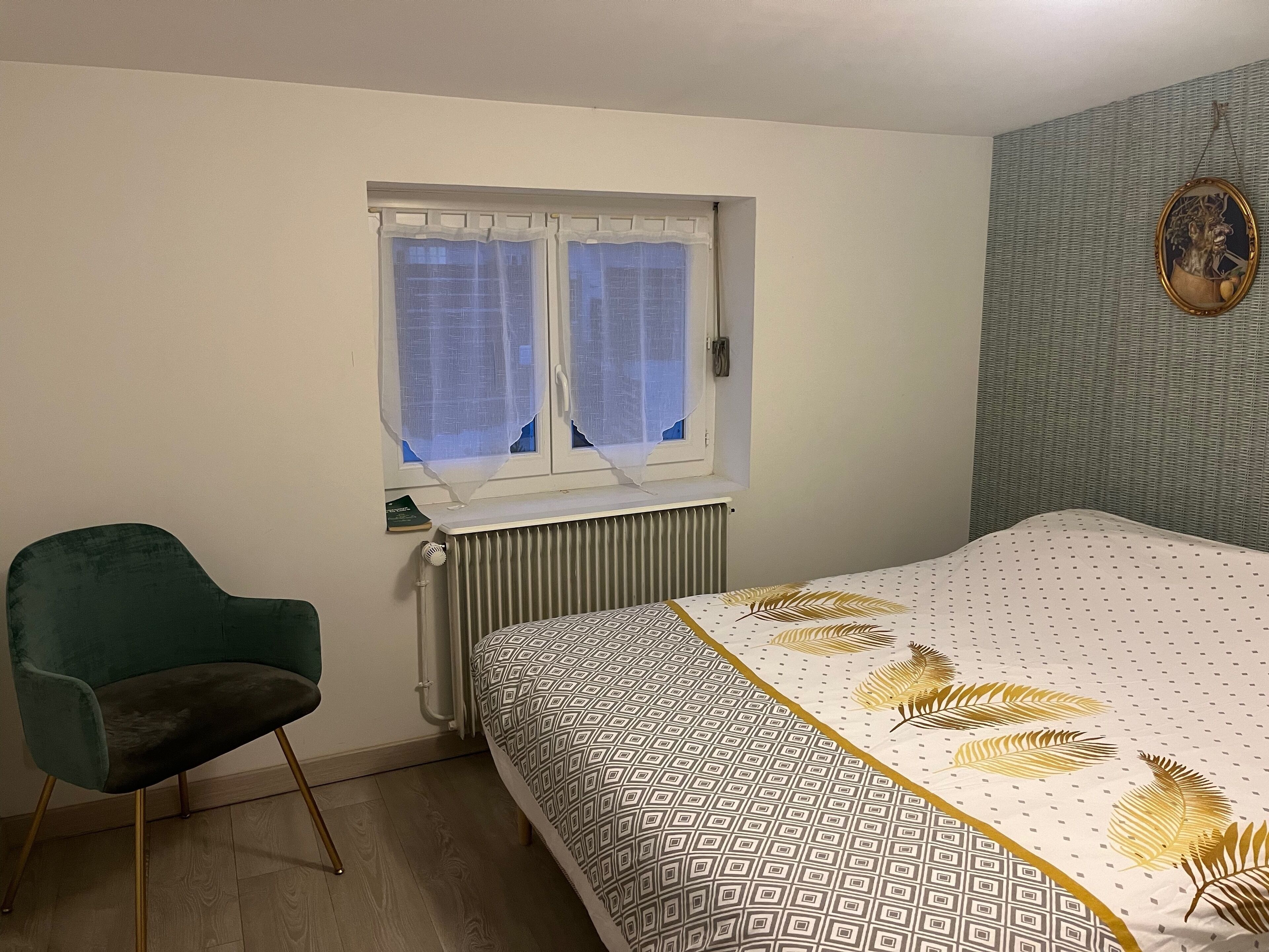 A bedroom with a bed, a radiator, a window with curtains, a chair, and a wall with a patterned wallpaper.