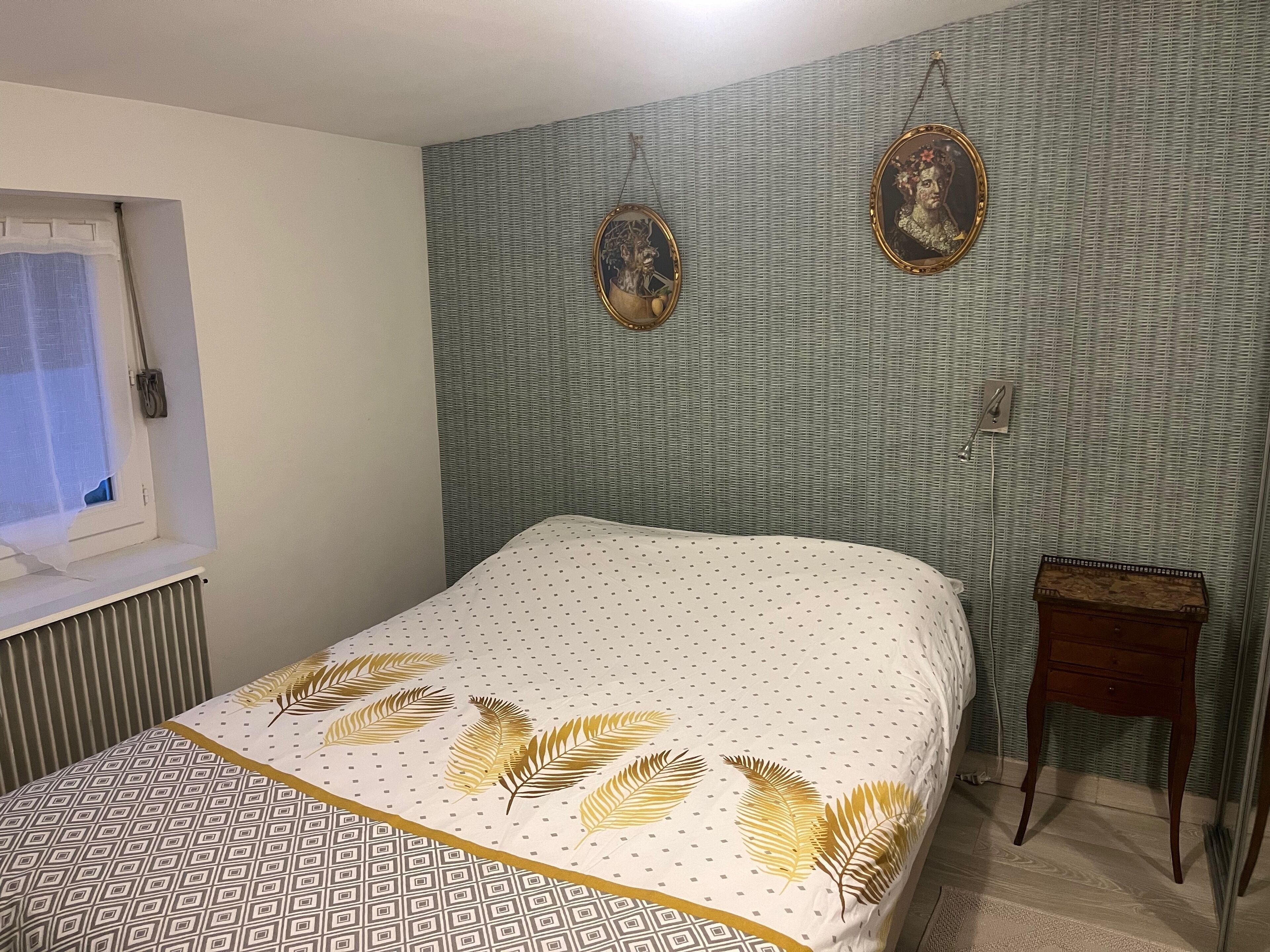 Ein Schlafzimmer mit Bett, Fenster, Heizkörper, einem kleinen Holztisch und zwei Bildern an der Wand.