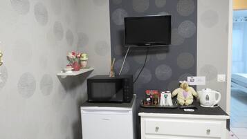 Camera d'albergo compatta con televisore a schermo piatto montato a parete, un piccolo frigorifero, una cassettiera bianca e uno scaffale con oggetti decorativi e un orsacchiotto.