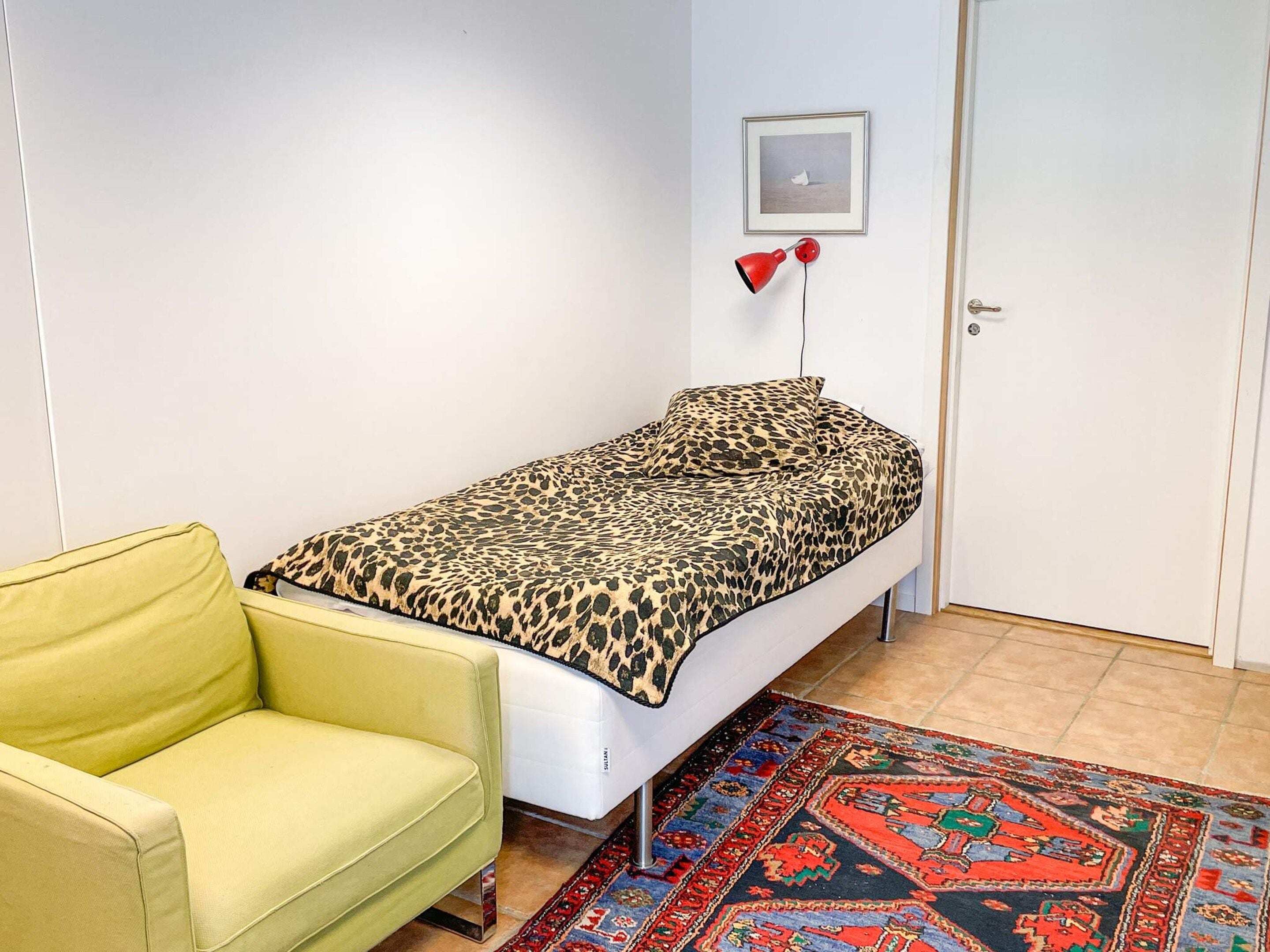 Una habitación pequeña con una cama individual, una silla amarilla, un tapete estampado y un cuadro en la pared.