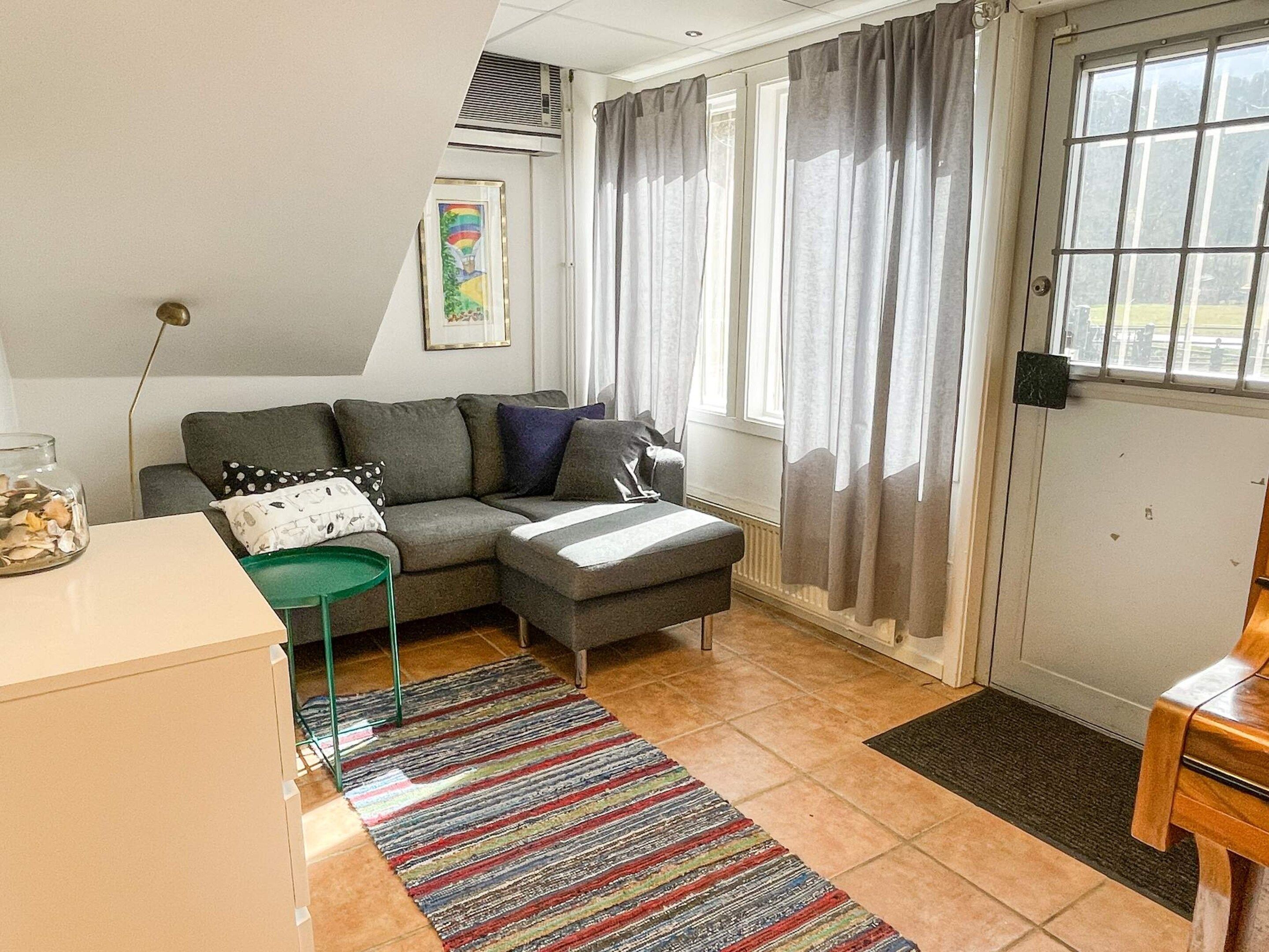 A living room with a sofa, a small table, a green chair, a colorful rug, and a window with curtains.