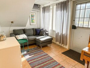 Una sala de estar con un sofá, una mesita, un sillón verde, un tapete colorido y una ventana con cortinas.