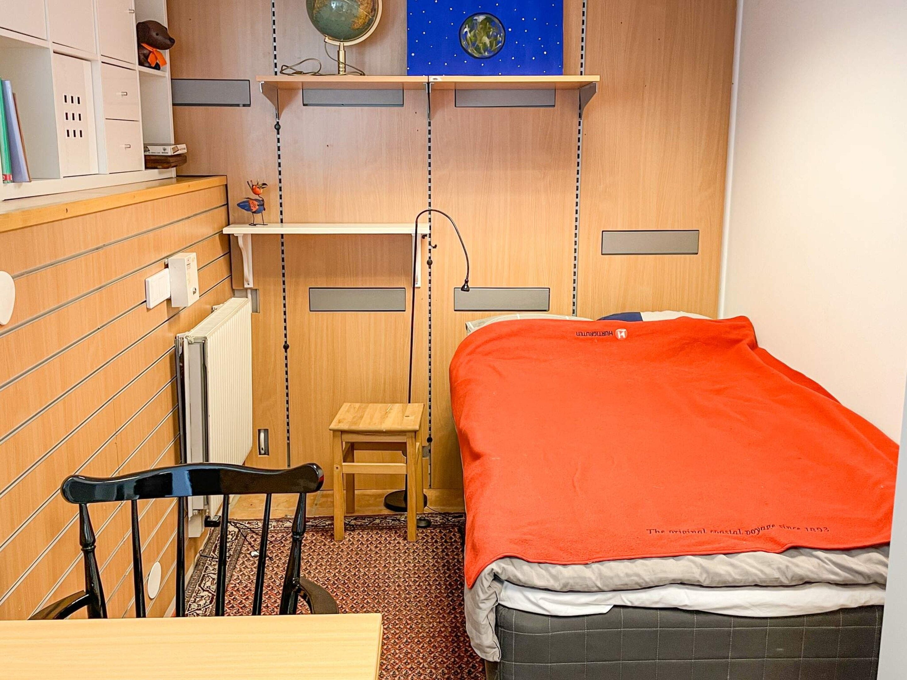 A compact room with a bed, a desk, a chair, and a small stool.