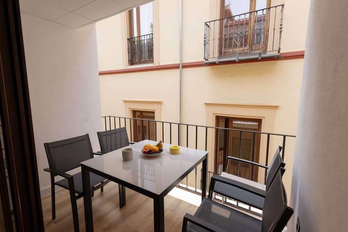 A small balcony with a table and chairs, a bowl of fruit, and a window with a view of buildings.