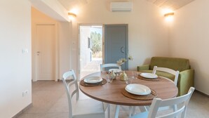 A dining area with a round wooden table, white chairs, and a green sofa. There is a door leading outside, and an air conditioning unit on the wall. - Lamia Flora con Piscina Privata (Latiano)