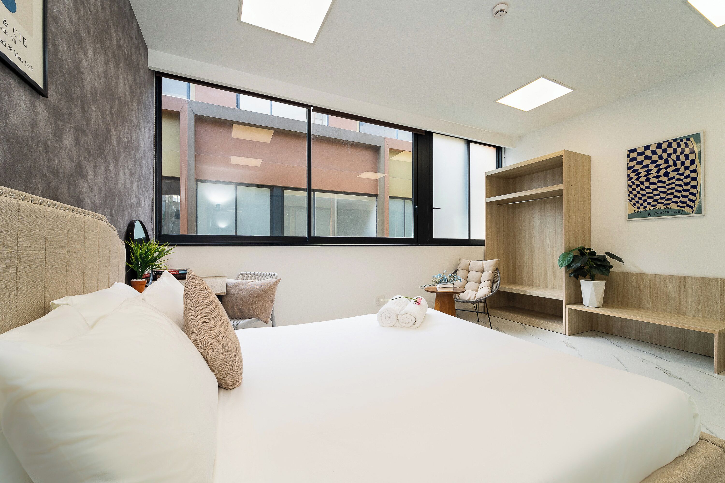 A modern hotel room with a large bed, a small seating area, a wooden wardrobe, and a large window offering a view of the outside.
