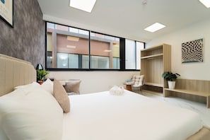 A modern hotel room with a large bed, a small seating area, a wooden wardrobe, and a large window offering a view of the outside. - The Global City - Jenny Residences (Ho Chi Minh City)