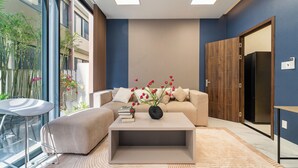A modern living room with a sofa, a coffee table, and a bookshelf. There is a large window with a view of plants and a balcony. - The Global City - Jenny Residences (Ho Chi Minh City)