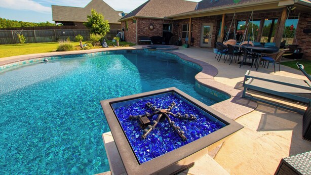 Una piscina con una fuente de fuego de mosaicos, mobiliario exterior y una casa de ladrillo al fondo.