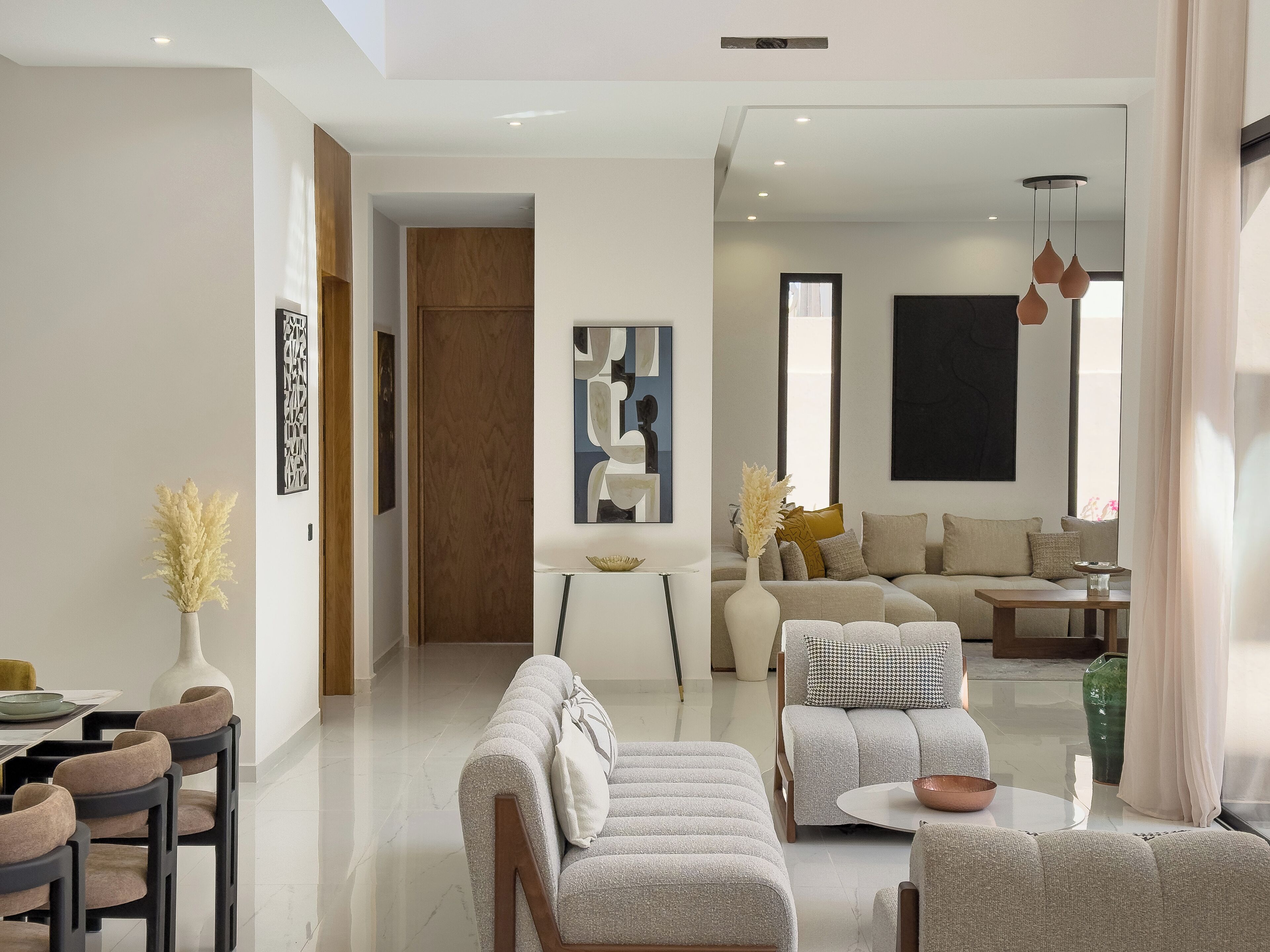 A modern living room with a sofa set, a dining area with chairs, and a coffee table. There is a vase with dried plants on the dining table.