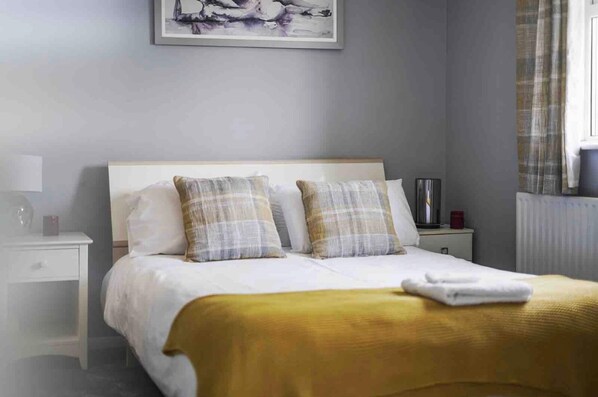 Una cama bien hecha con un edredón amarillo y dos almohadas, una mesita de noche con una lámpara y un cuadro en la pared.
