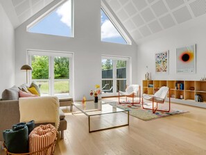 A spacious living room with a large glass coffee table, a sofa, and two chairs. There are large windows providing natural light and a view of greenery outside.