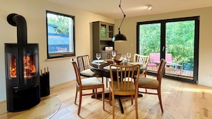 A dining area with a wood-burning stove, a round table with chairs, and a view of greenery through large windows.