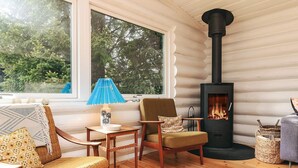 A cozy cabin interior with a wooden floor, a black wood stove, and a view of trees through large windows.