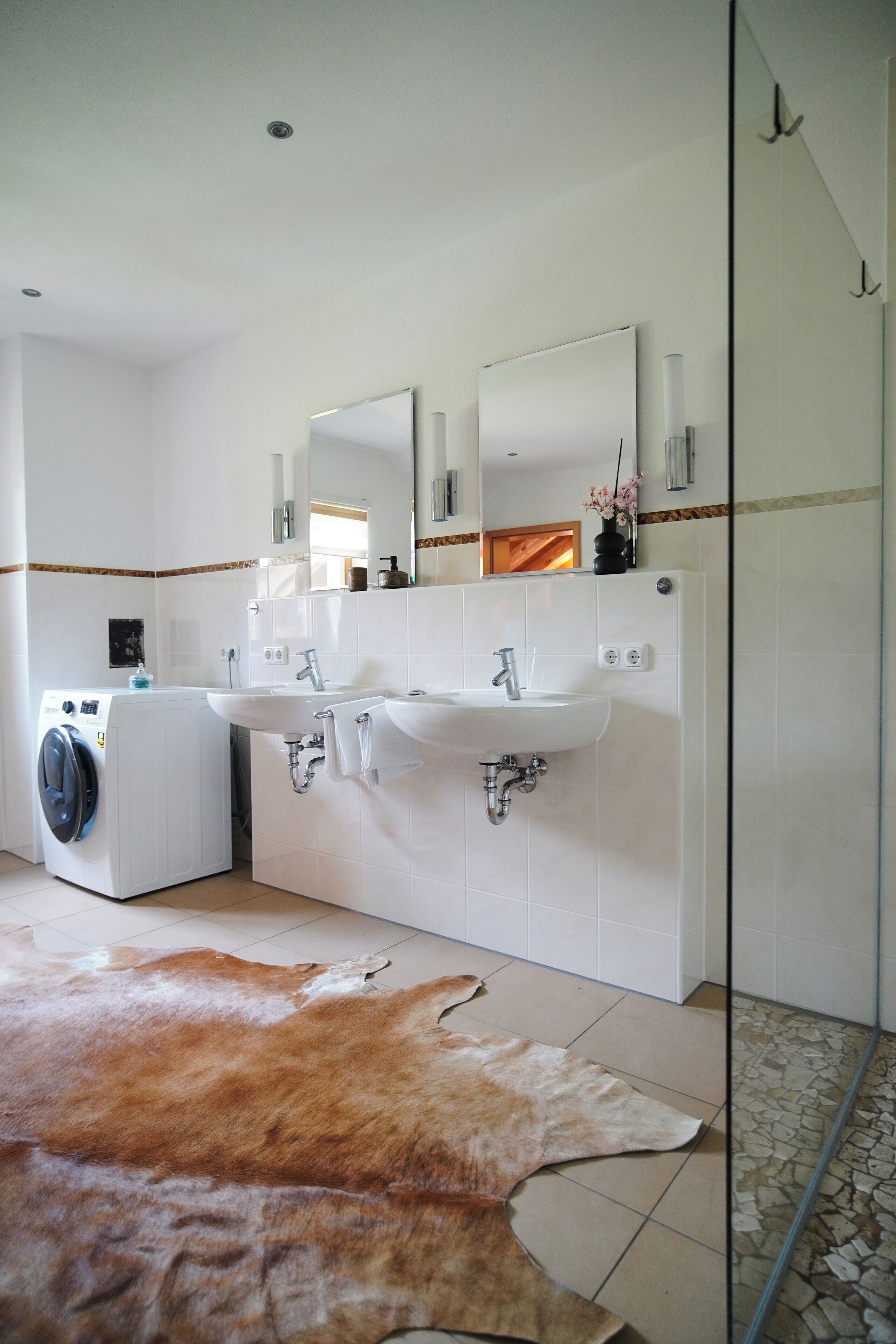 Ein modernes Badezimmer mit einem großen Spiegel, zwei Waschbecken, einer Waschmaschine und einer Duschkabine aus Glas.
