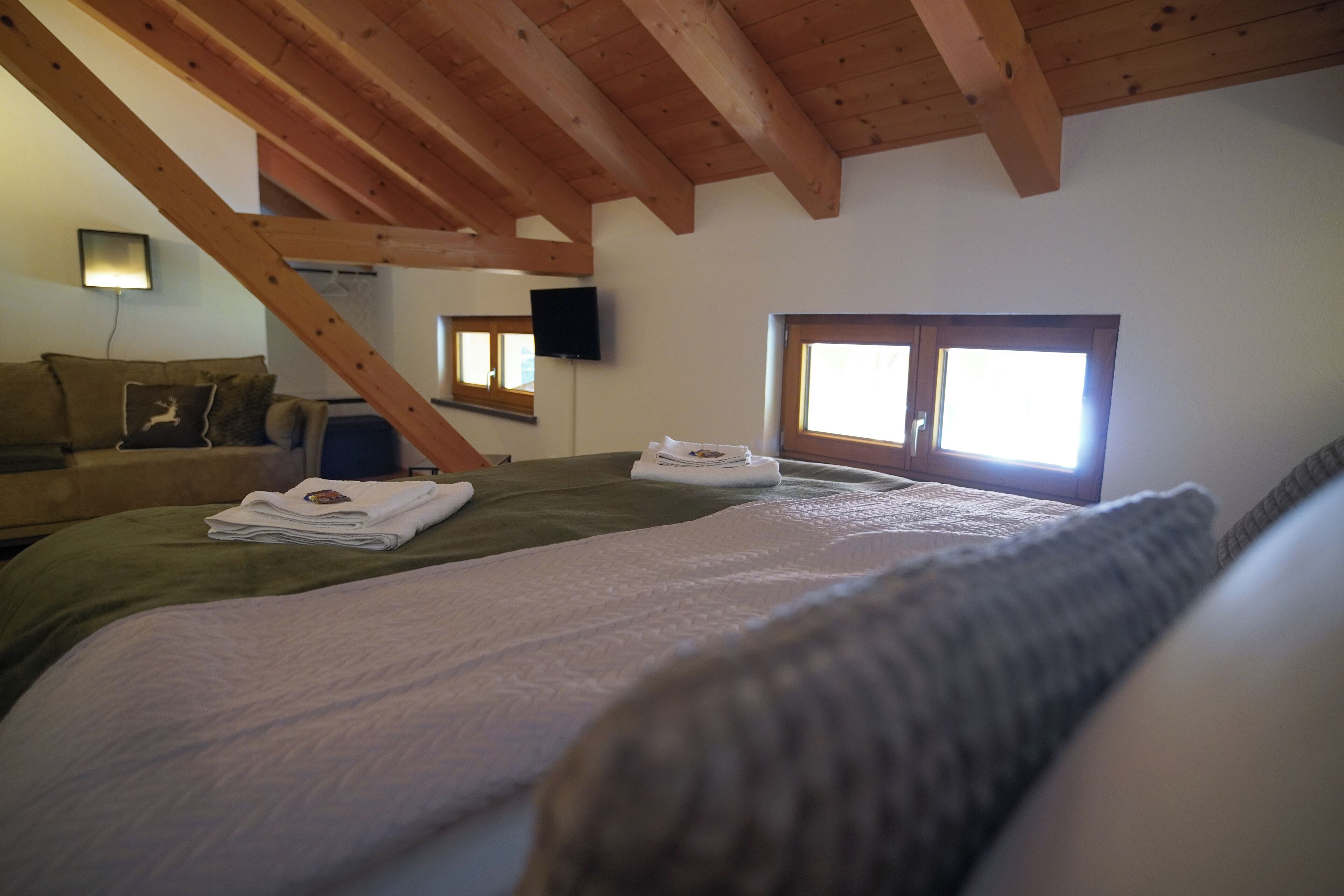 A bedroom with a wooden ceiling, two beds with neatly folded towels, a green blanket, a window, and a wall-mounted TV.