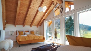 A bedroom with a wooden ceiling, a large bed with orange bedding, a fur rug, and a view of a green landscape through large windows. - Alpen Lodges Gindels - Space for 10 - (Rettenberg)