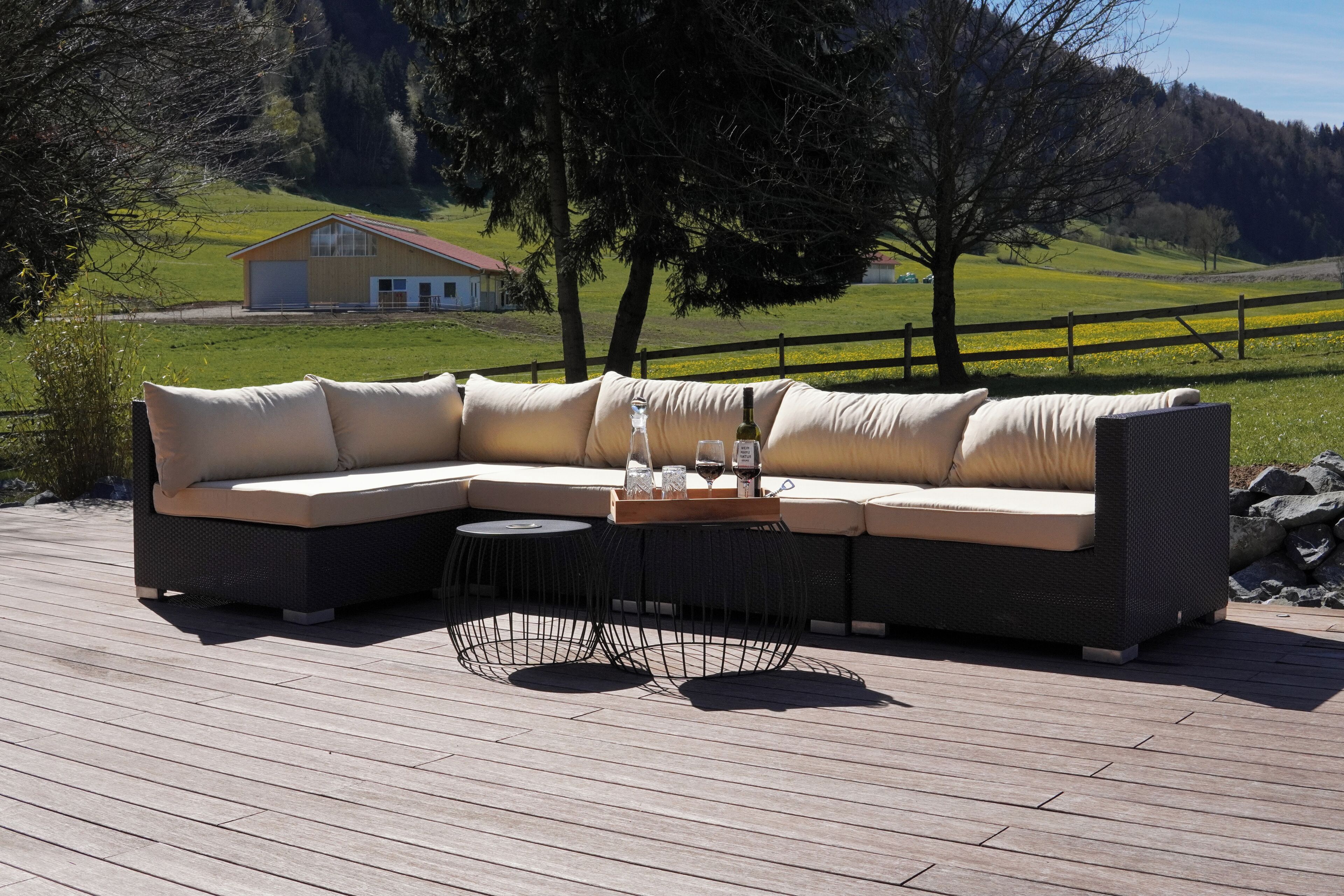 A sectional sofa with cushions on a wooden deck, a small round table with a tray holding glasses and a bottle, and a house in the background.