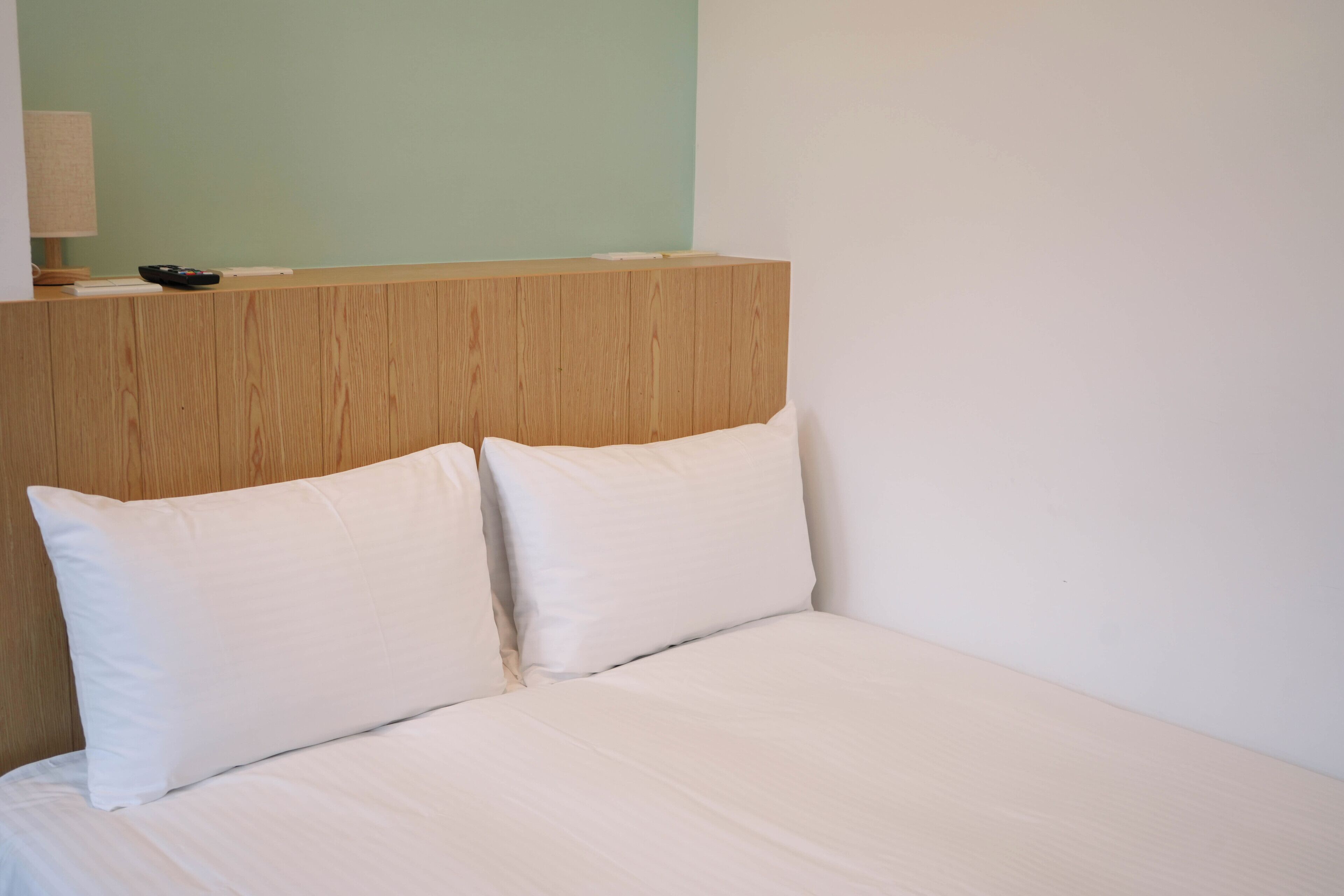 A hotel room with a wooden headboard, two white pillows, and a bedside table with a lamp and remote control.
