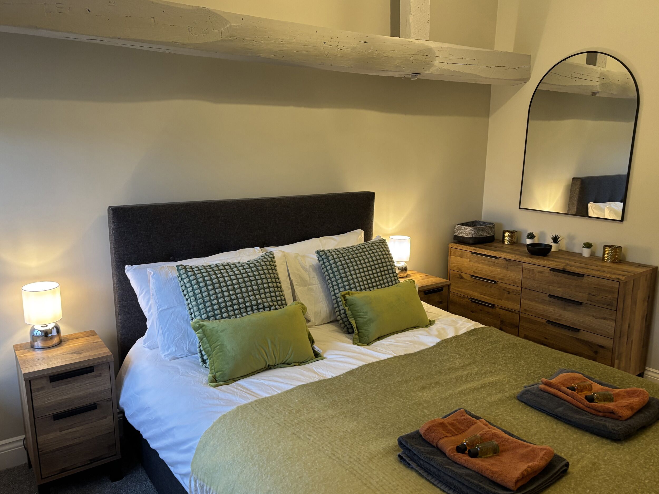 A neatly made bed with pillows, a bedside lamp, a wooden dresser, and a mirror.