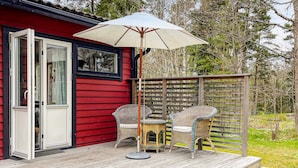 A wooden deck with wicker chairs and a table, a red building with a glass door, and a wooden fence in a garden setting. - 4 Star Holiday Home in Norrtalje (Norrtälje)