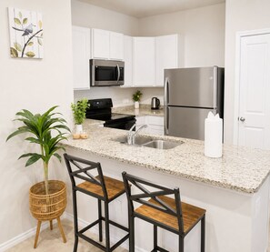 A kitchen with white cabinets, a stainless steel refrigerator, a microwave, a sink, and a bar stool area.