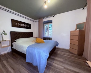 A bedroom with a bed, a nightstand, a dresser, a window, and a wall art piece.