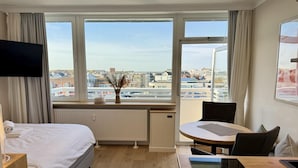 A hotel room with a bed, a dining table, a chair, a TV, a window with a view of buildings, and a radiator. - Seeluft - Modern 1-room apartment with balcony and sea view, parking space, WLAN. (Sylt)