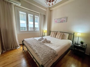 A neatly made bed with a white comforter and pillows, a bedside table with a lamp, and a window with curtains.