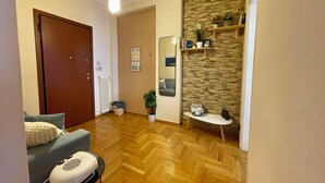 A living room with a wooden herringbone floor, a grey sofa, a white armchair, a small table with a basket, and a wall-mounted shelf with decorative items.