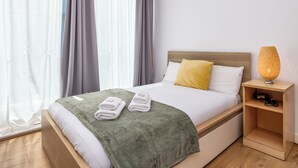 A neatly made bed with a green blanket and two white towels, a wooden nightstand with a lamp, and a window with grey curtains. - 3 Bed in Dublin city centre 10min to Spire (Dublin 1)