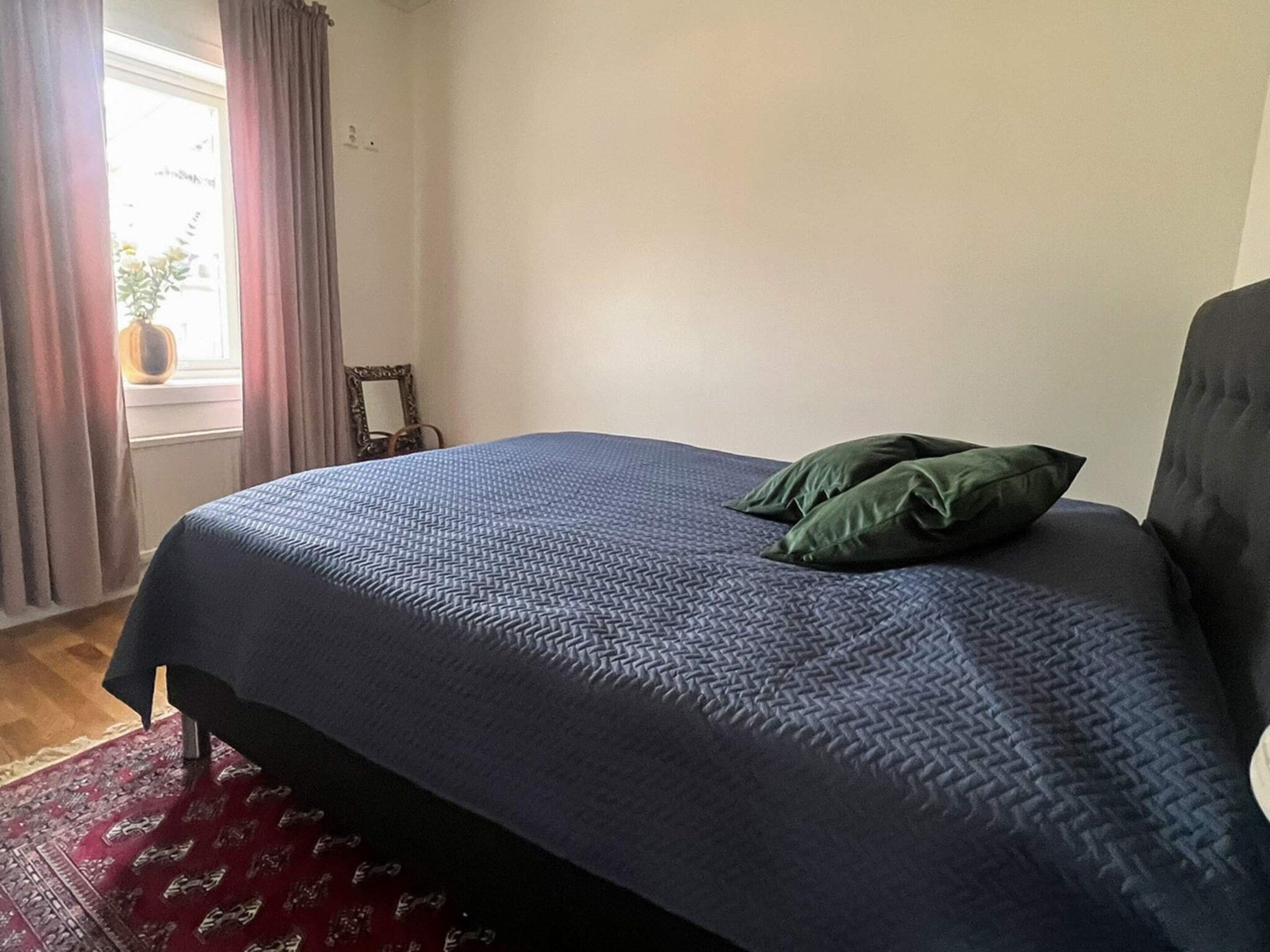 A room with a bed, a window with curtains, a small table with a mirror, and a red patterned rug.