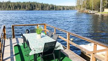 A wooden deck with a table and chairs, overlooking a lake with a forested shoreline.